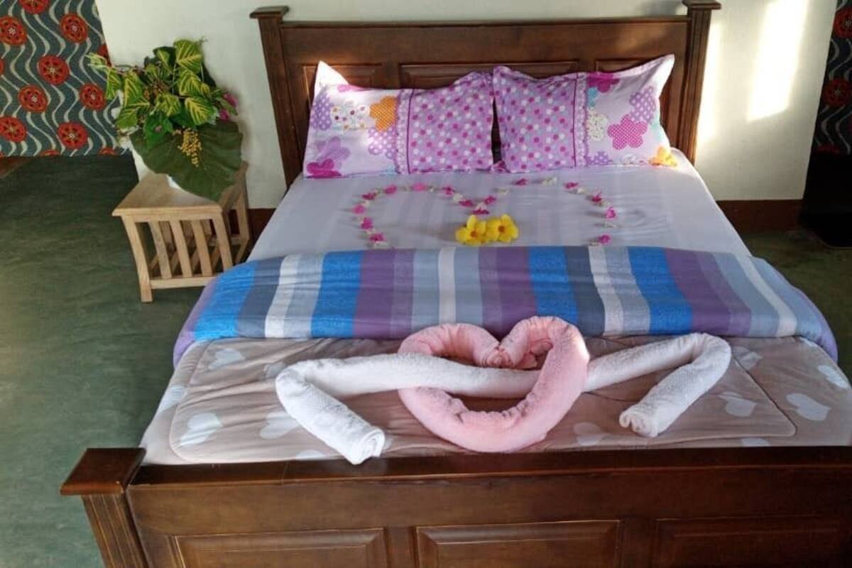 A neatly made bed with a heart-shaped towel arrangement, a decorative pillow, and a small plant on a wooden nightstand.