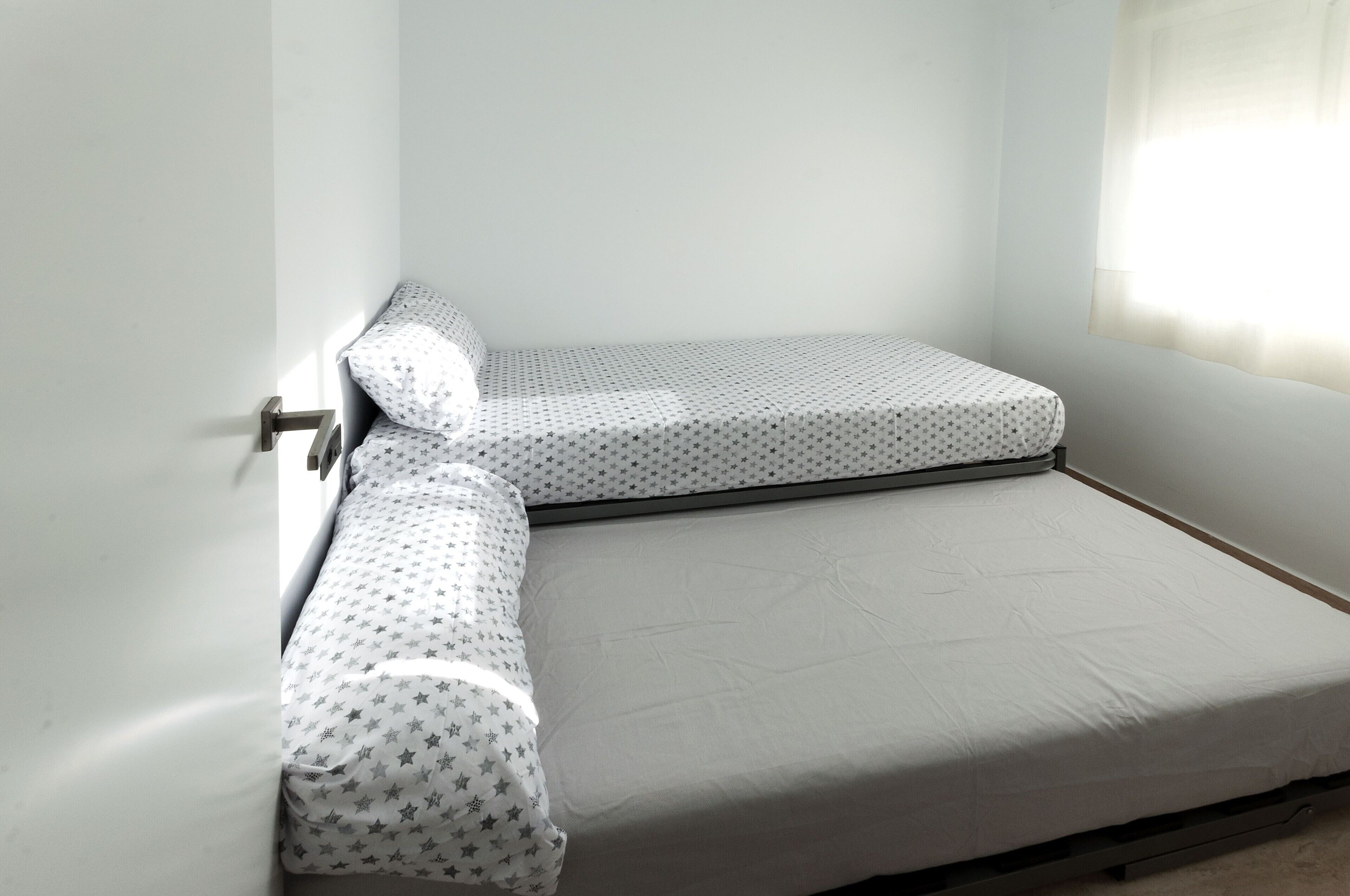 Una habitación pequeña con una cama individual, colcha blanca con estampado de estrellas y colchón blanco liso.