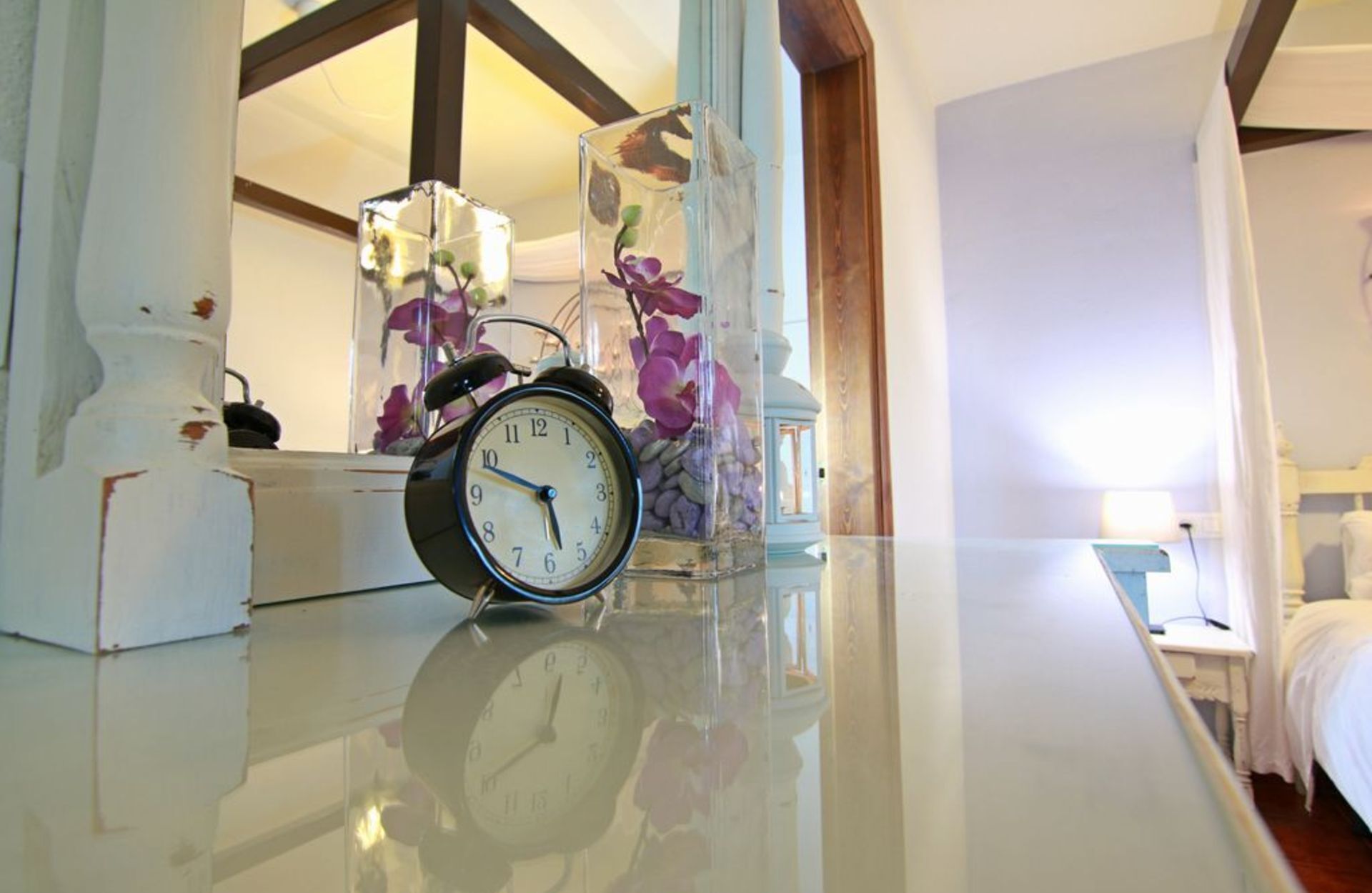 Un reloj sobre una mesa, con un jarrón de flores al fondo.