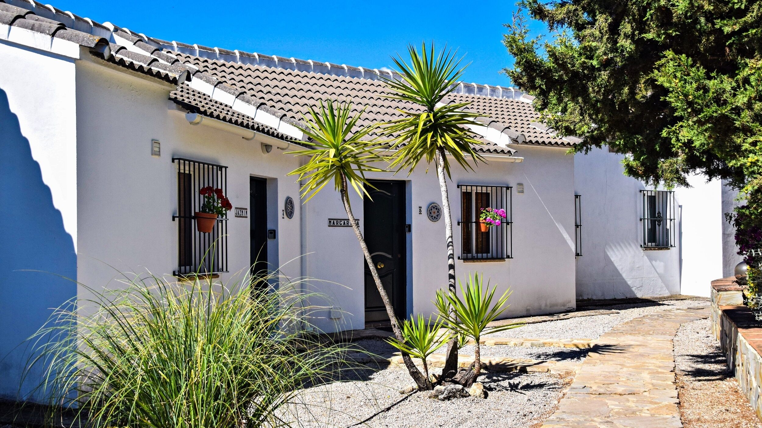 A white building with a tiled roof, a door, and windows with flower boxes. There are plants in front of the building.