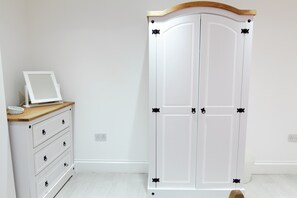Une armoire blanche avec des accessoires noirs, une commode en bois avec des tiroirs et un mur blanc avec une prise de courant.