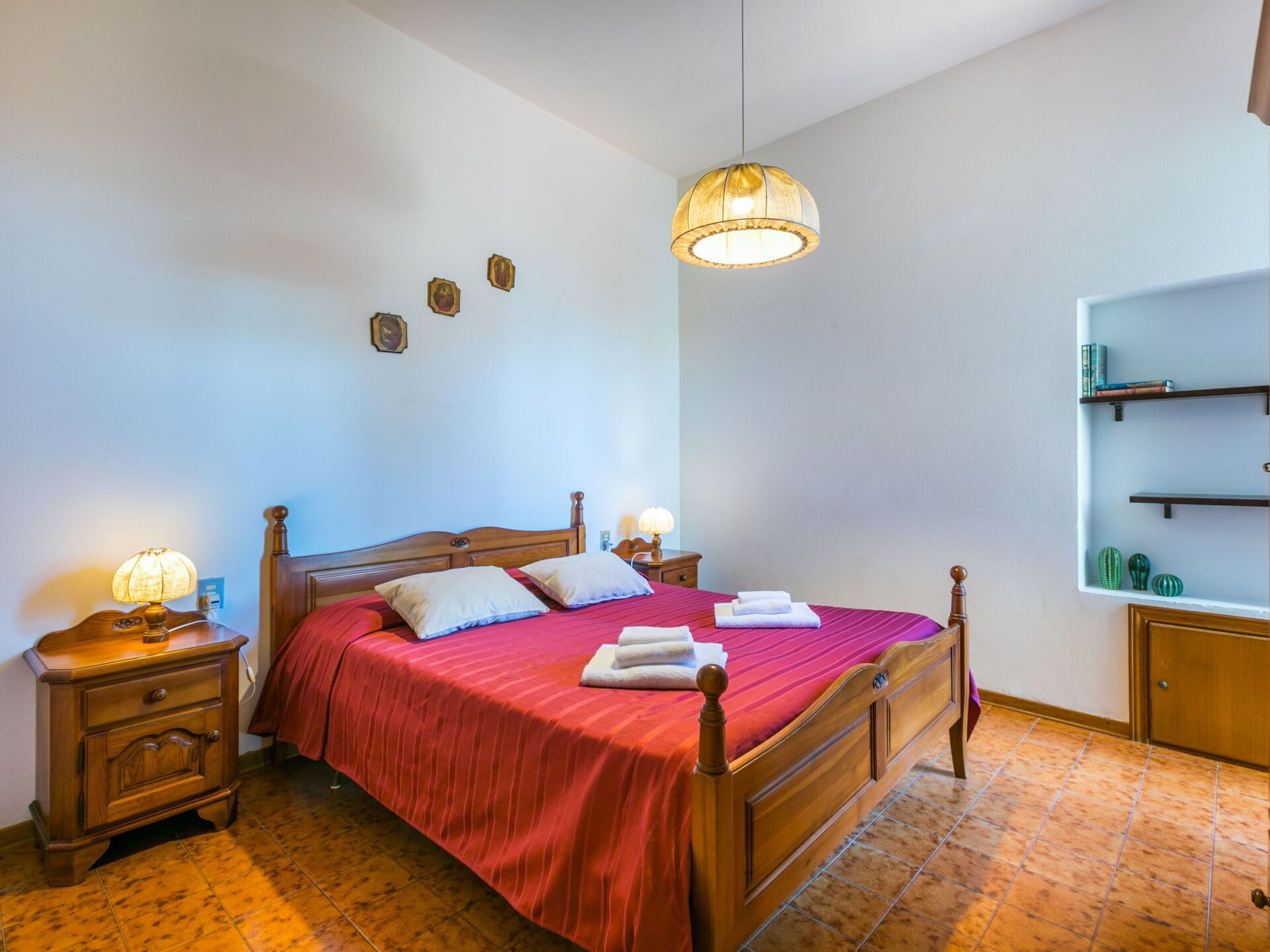 Een netjes ingerichte slaapkamer met een houten bed, nachtlampjes, een hanglamp en een klein plankje met boeken en decoratieve objecten.