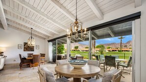 A spacious patio area with a round wooden table and chairs, a hanging light fixture, and a view of a golf course and mountains. - LQ001 - 3 bedroom 3 bath with pool (La Quinta)