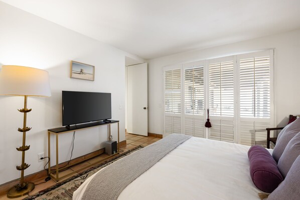 A hotel room with a bed, a TV on a stand, a lamp, and a window with blinds.