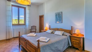 Une chambre à coucher avec un lit en bois, deux tables de chevet avec des lampes, une chaise, un radiateur et une fenêtre avec des rideaux.