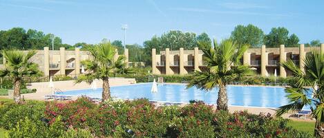 A pool area with palm trees, a clear sky, and a building with balconies in the background.