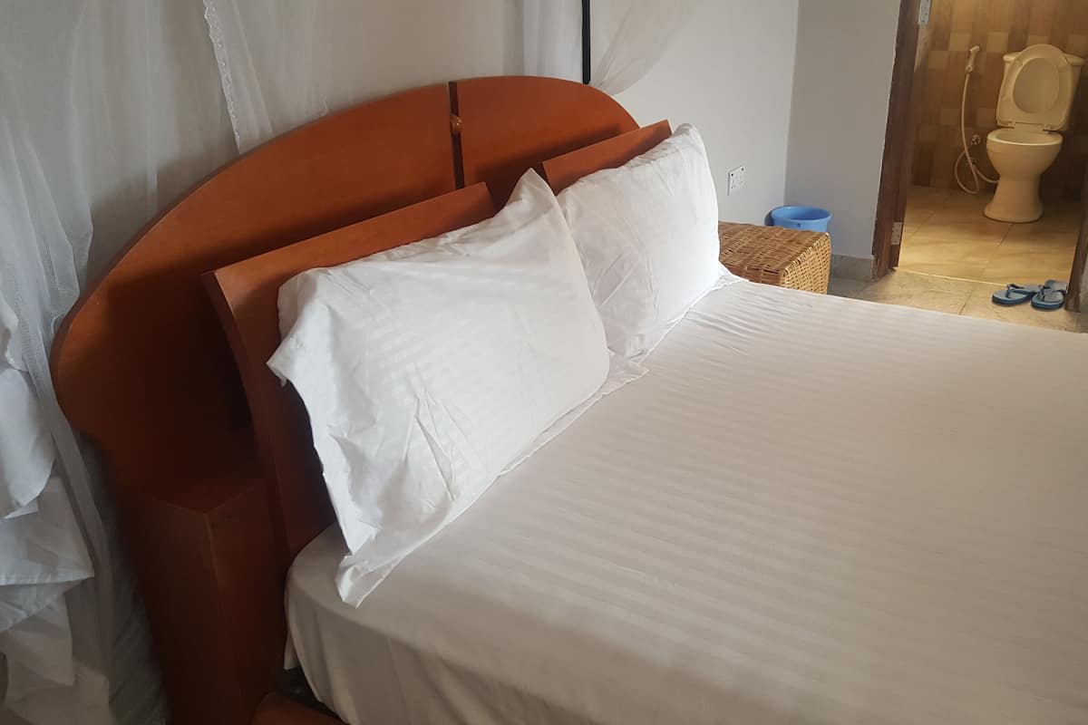 A bed with white linens, a wooden headboard, and a view of a bathroom with a toilet and a blue bucket.