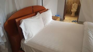 A bed with white linens, a wooden headboard, and a view of a bathroom with a toilet and a blue bucket.