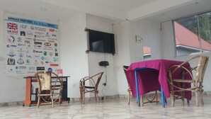 A room with a tiled floor, a television mounted on the wall, a table with a pink tablecloth, and several wicker chairs. - Guest House in Arusha -Nanofilter Family (Arusha)