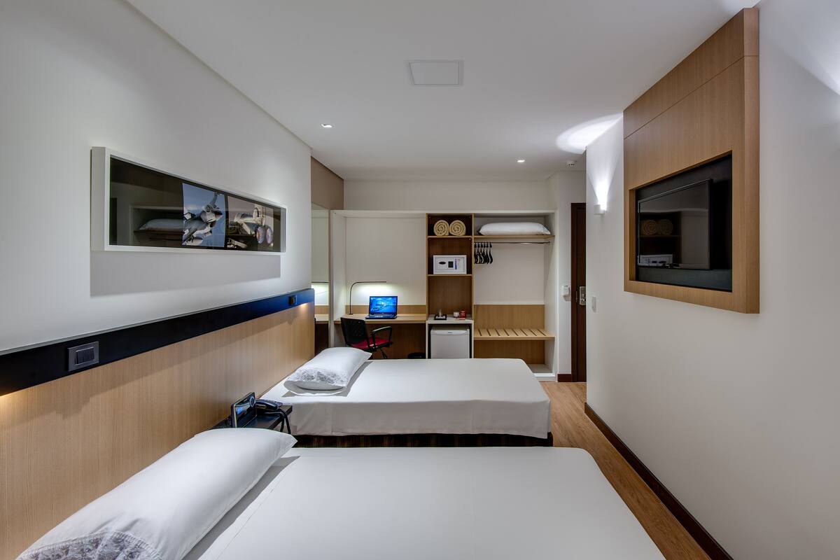 Une chambre d’hôtel avec deux lits, un bureau avec un ordinateur, un petit réfrigérateur et un miroir au mur.