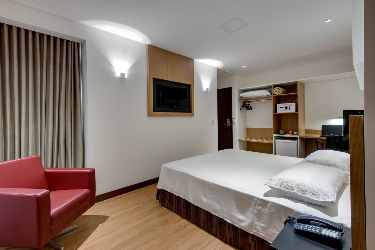 Une chambre d’hôtel avec un lit, une chaise rouge, un téléviseur fixé au mur et un petit bureau avec un téléphone.