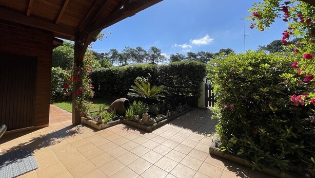 Un patio cubierto con piso de baldosas, techo de madera y vistas a un jardín con plantas variadas y un seto.