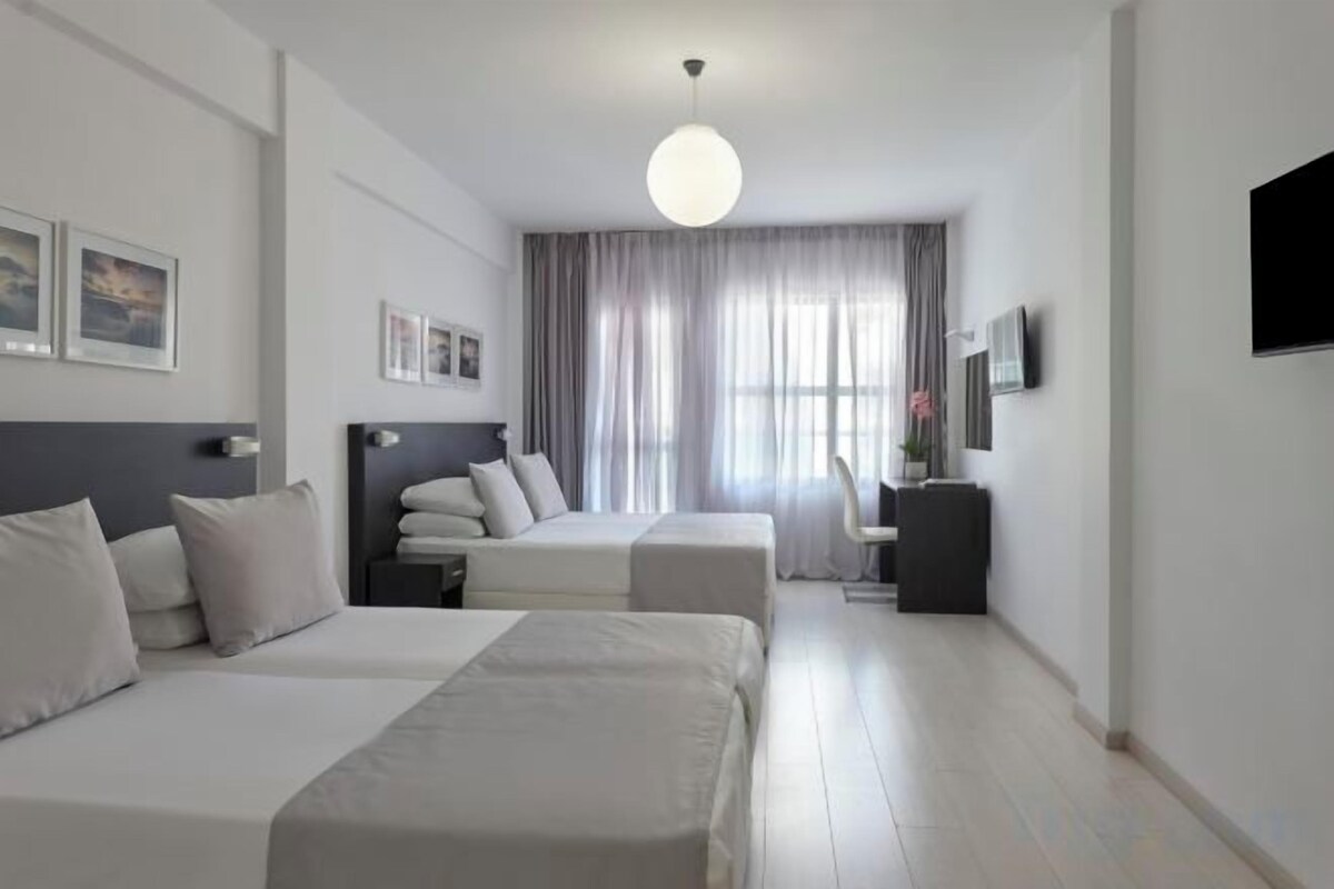 Habitación de hotel con dos camas, un escritorio, una silla, un televisor y un ventanal con cortinas.