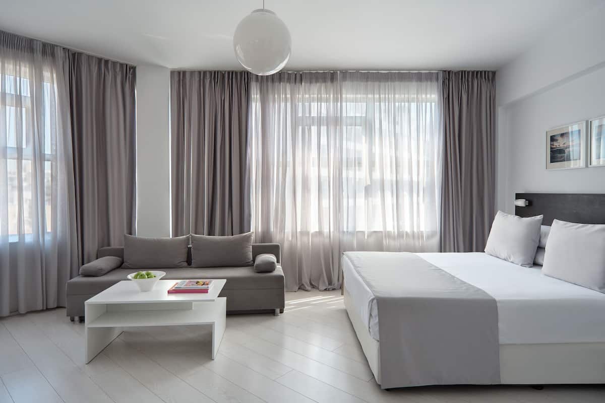 A modern hotel room with a large bed, a sofa, a coffee table, and sheer curtains.