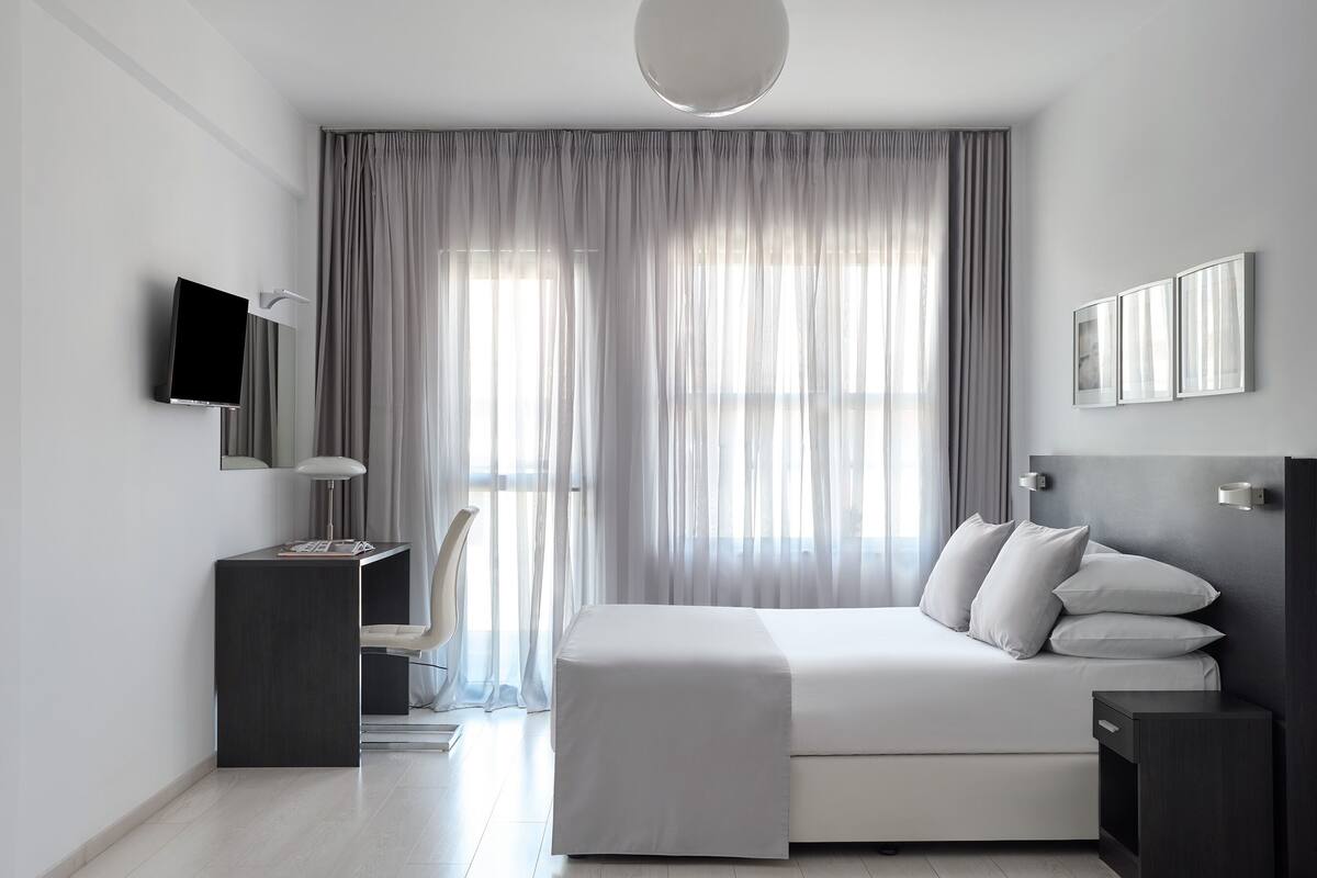 A modern bedroom with a bed, bedside tables, a TV, and a large window with curtains.