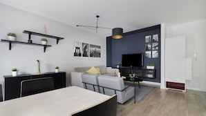 Un salon moderne avec un canapé, une table basse et un téléviseur. On y trouve des étagères avec des plantes et un coin repas avec des chaises.
