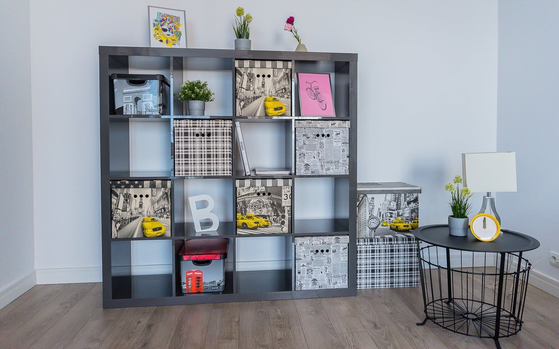 A modern room with a black shelving unit displaying various framed artworks and a small round table with a lamp and flowers.
