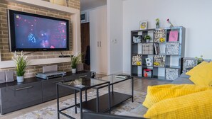 A modern living room with a flat-screen TV, a glass coffee table, a sofa with yellow cushions, and a bookshelf with various items. - Easy Rent Apartments - SKY (Lublin)