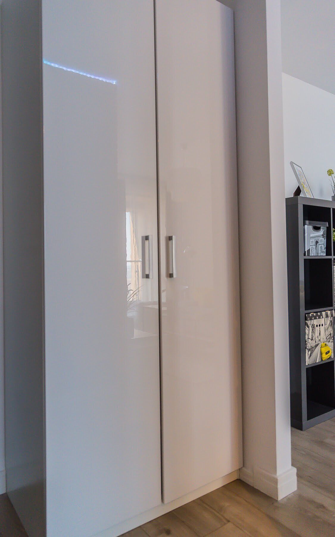 A white wardrobe with two doors and a handle, located in a room with wooden flooring and a black shelf with decorative items.