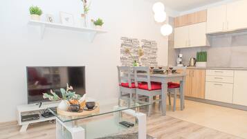 Una cocina moderna con armarios de madera clara, una zona de comedor con una mesa de vidrio y sillas rojas, y una sala de estar con un televisor de pantalla plana y una mesa de centro de vidrio.