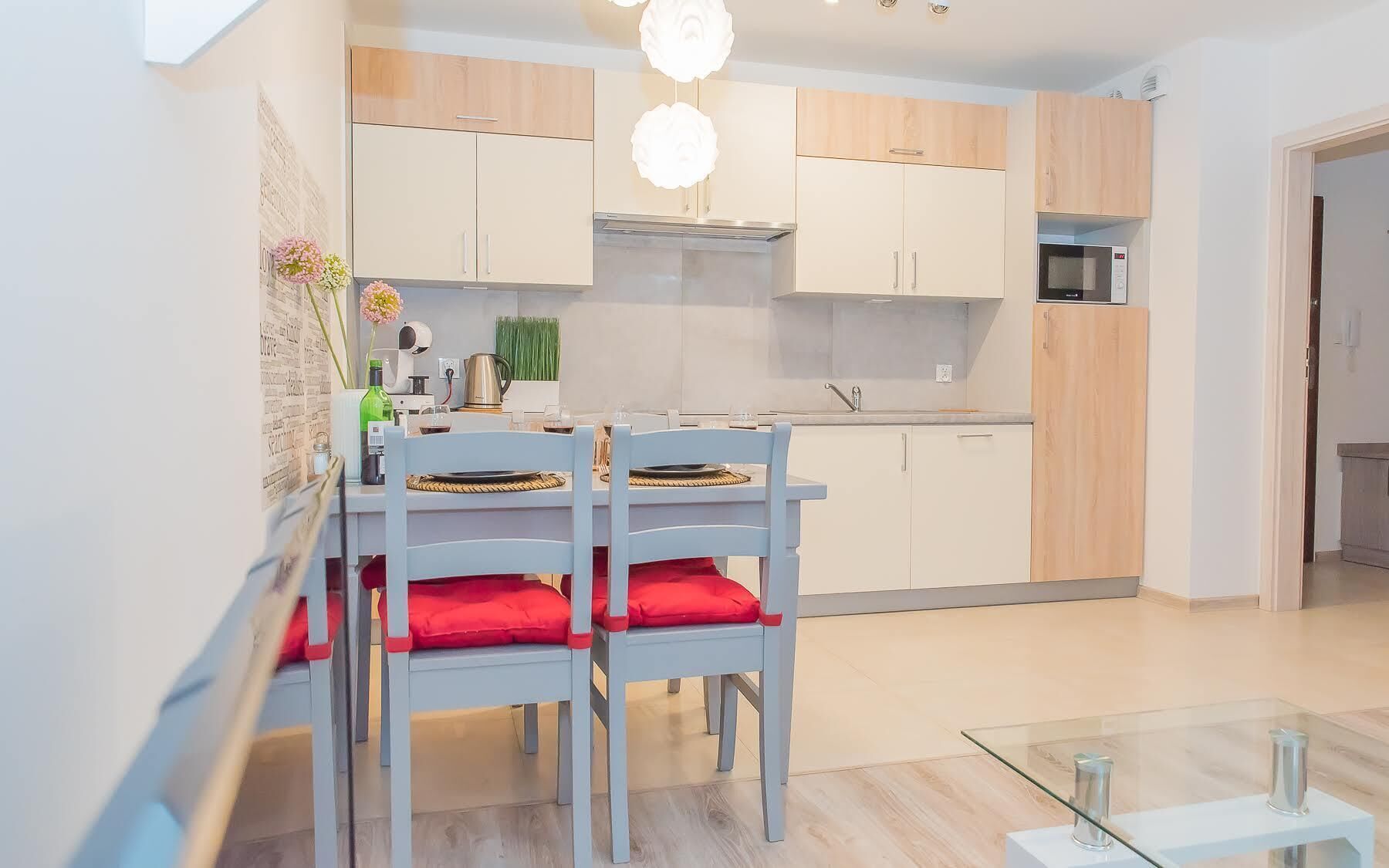 A modern kitchen with light wood cabinets, a stainless steel range hood, and a glass dining table with two chairs.