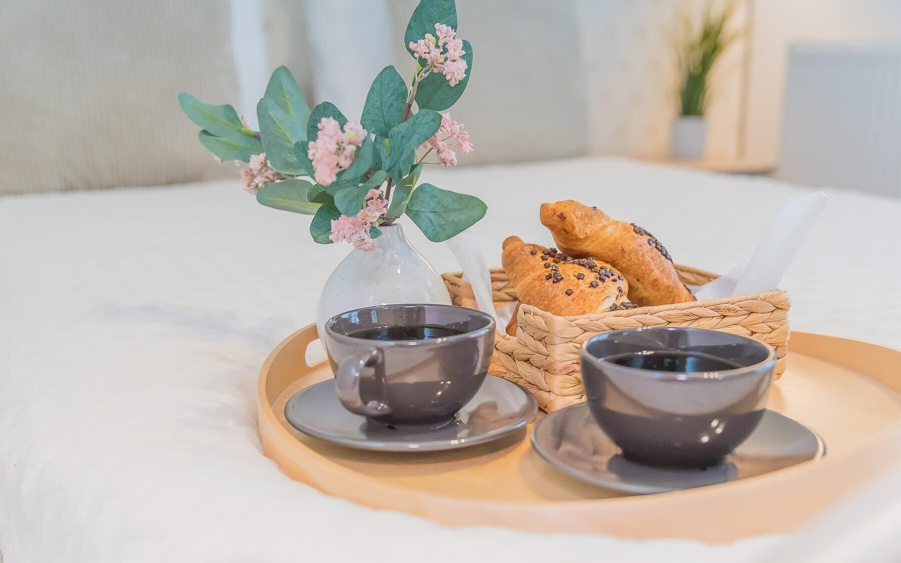 A breakfast tray with two cups of coffee, a vase with pink flowers, and a basket of croissants on a bed.