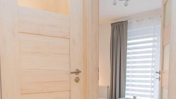 Un pasillo que conduce a un dormitorio con una cama, un ventanal con persianas y una puerta con interruptor de luz.