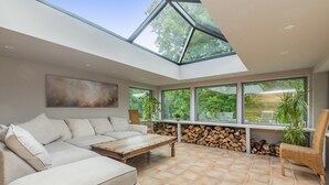 A modern living room with a large sectional sofa, a wooden coffee table, and a wicker chair. There are large windows providing natural light and views of greenery outside. - Poundgate Park Holiday House 8 Bedroom in 2 adjoining properties ,1h from London (Crowborough)