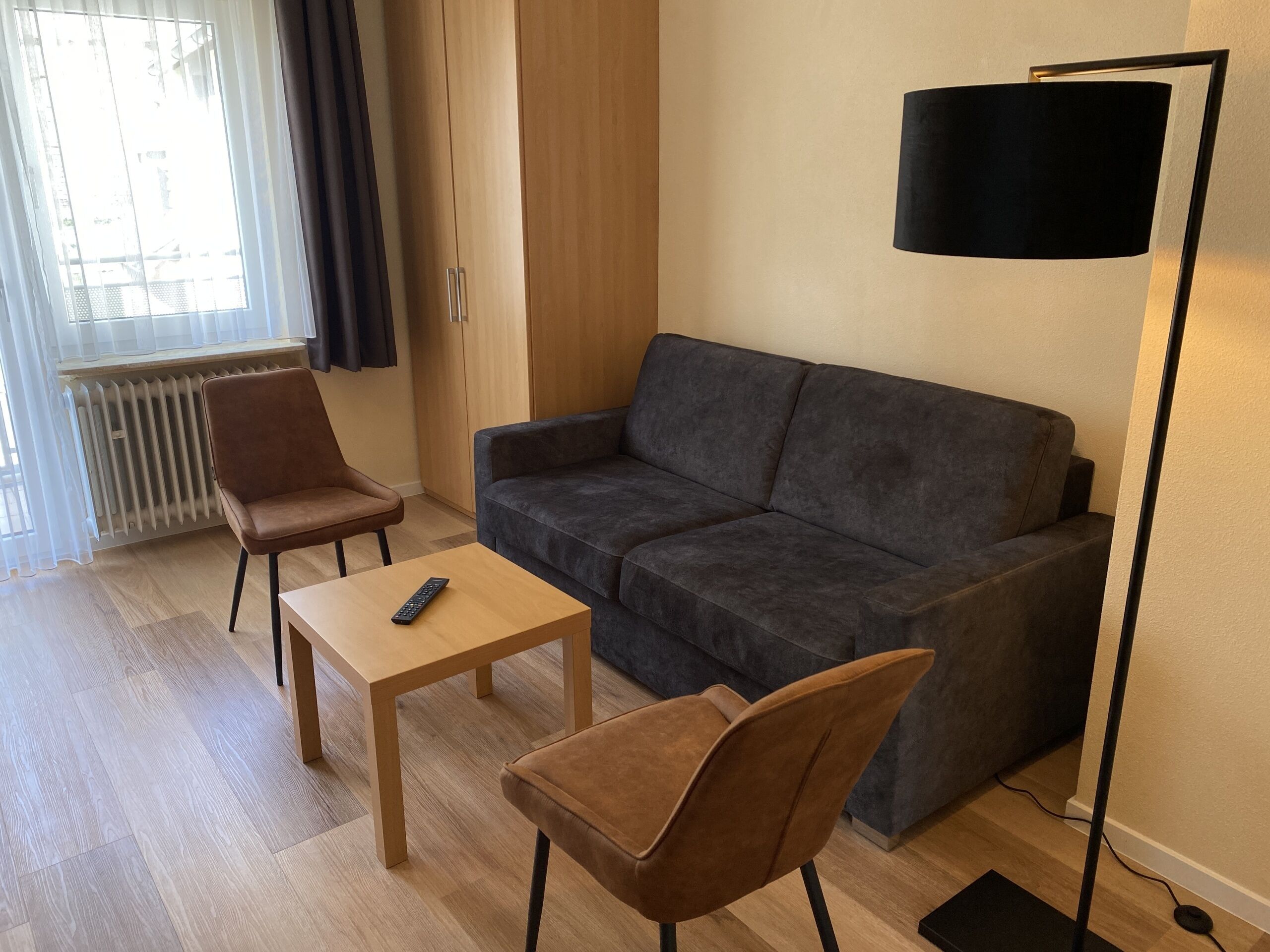 A modern living room with a grey sofa, two brown chairs, a small wooden table with a remote control, a floor lamp, and a radiator.