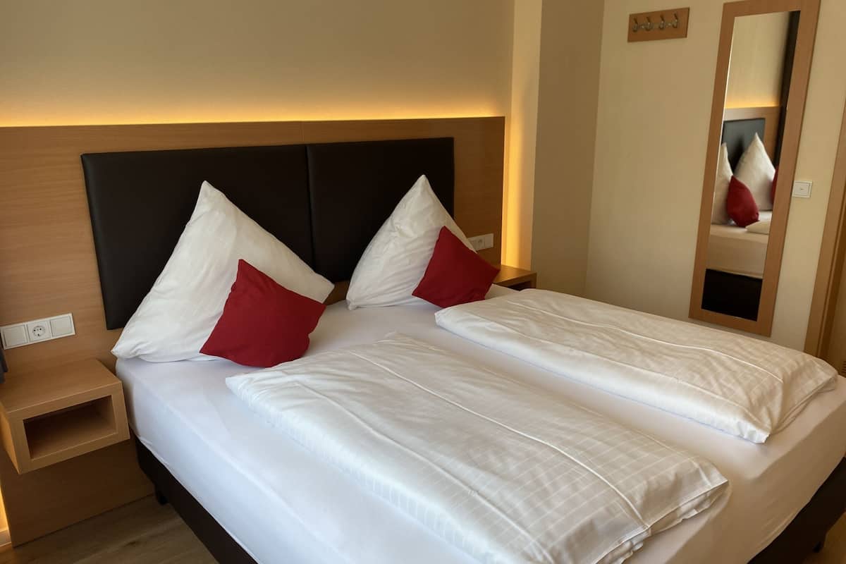 A double bed with white and red pillows, a wooden headboard, and a bedside table with a lamp.