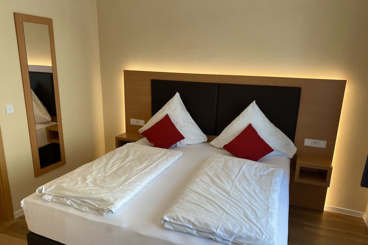 A double bed with white linens and red pillows, a wooden headboard, bedside tables, and a mirror on the wall.