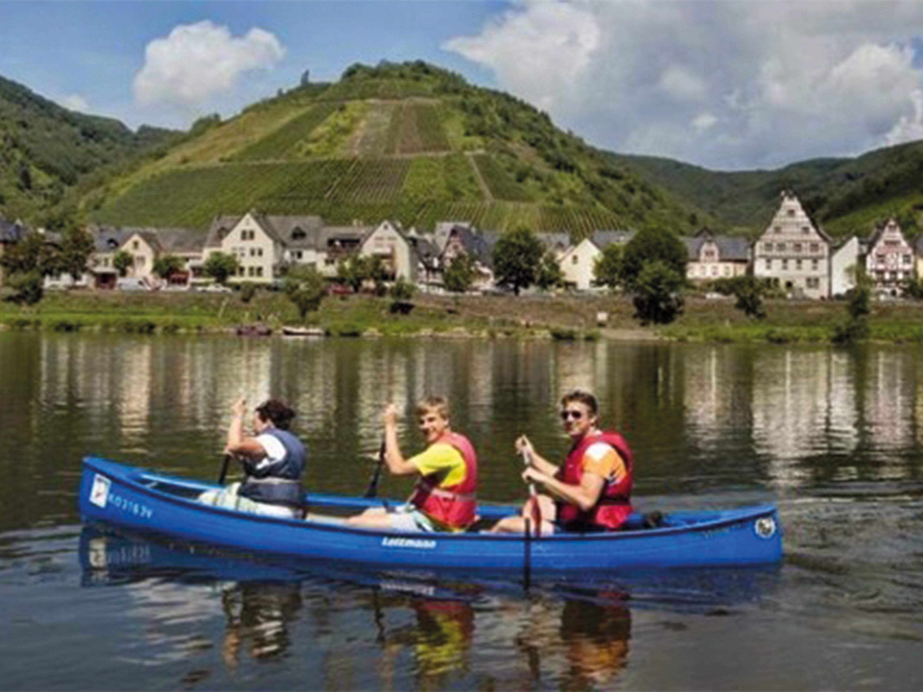Un canoë bleu sur un lac paisible, avec des personnes qui pagayent, entouré de maisons et d’une colline en arrière-plan.