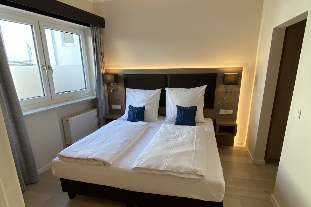 A neatly made bed with white linens and two blue pillows, a bedside table with a lamp, and a window with curtains.