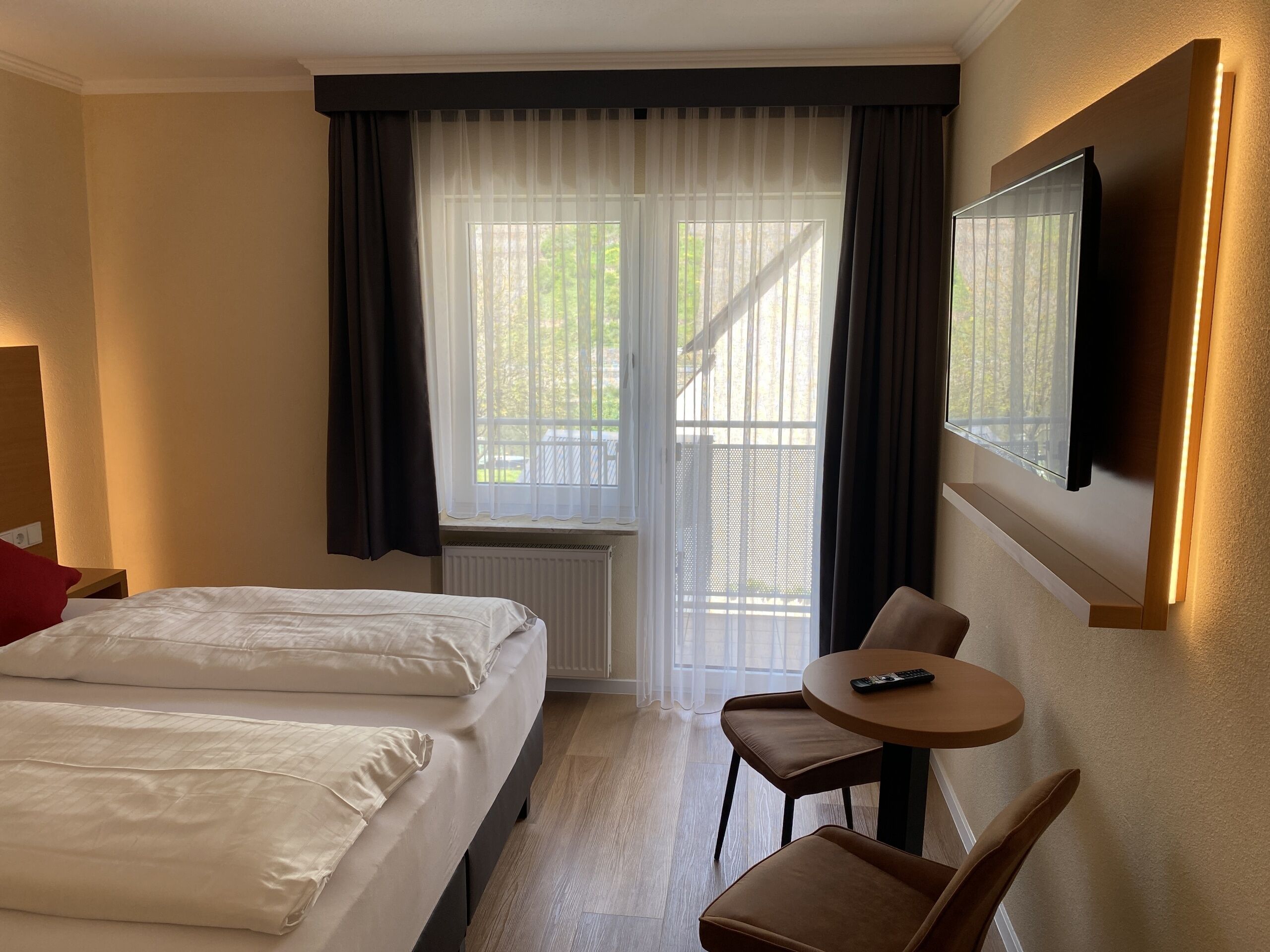 A hotel room with two beds, a small table with a chair, a TV mounted on the wall, and a window with curtains.
