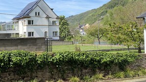 A white two-story house with a sloped roof, a stone wall with ivy, and a view of a river and hills in the background. - Moseltel Ernst/cochem (Ernst)