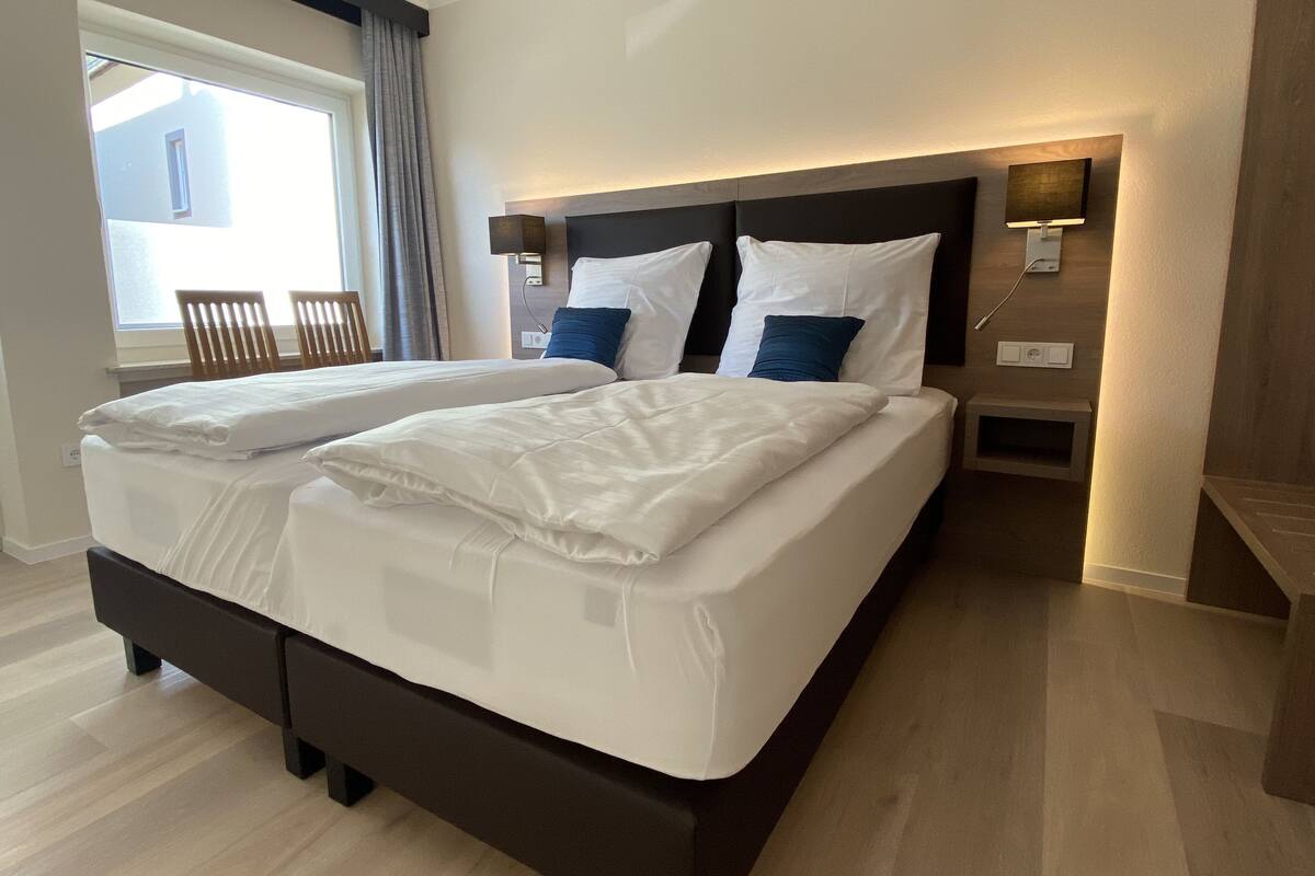 A double bed with white linens, a headboard with integrated lighting, a bedside table with a lamp, and a window with curtains.
