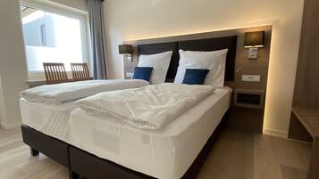 Una cama doble con sábanas blancas, cabecera con iluminación integrada, mesita de noche con lámpara y ventana con cortinas.