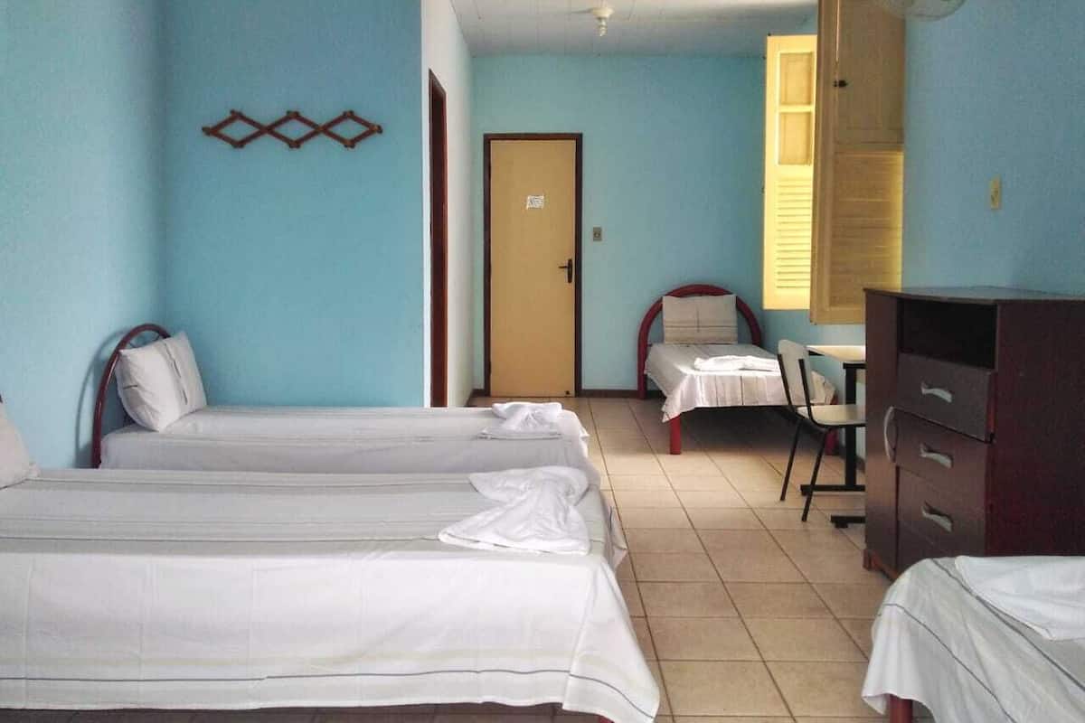 Una habitación con tres camas individuales, un escritorio con silla, un armario y un ventilador de pared.