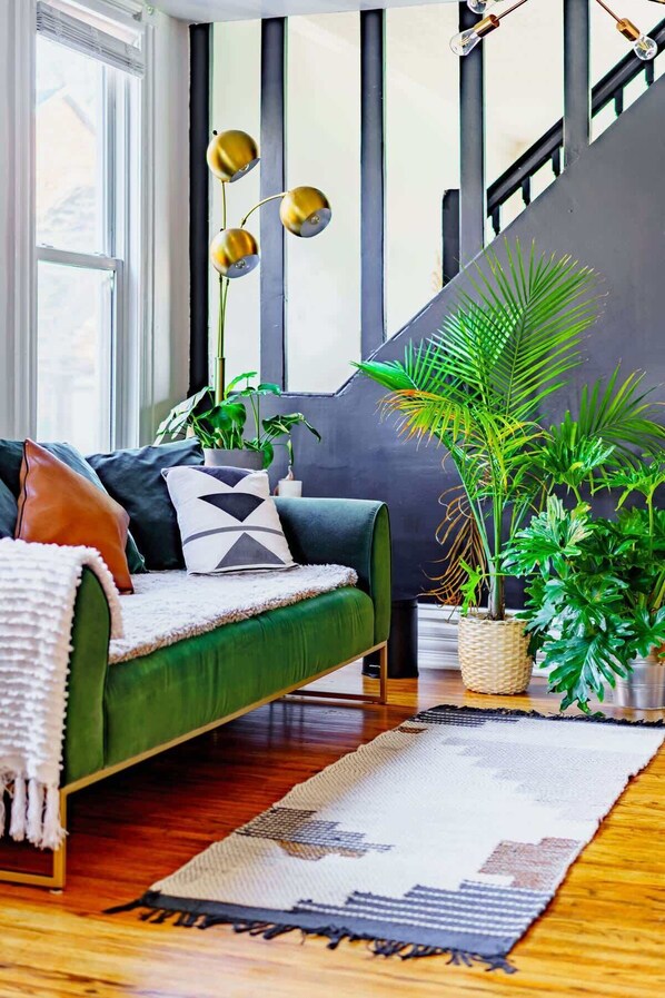Una sala de estar moderna con un sofá verde, varias macetas con plantas y una escalera al fondo.