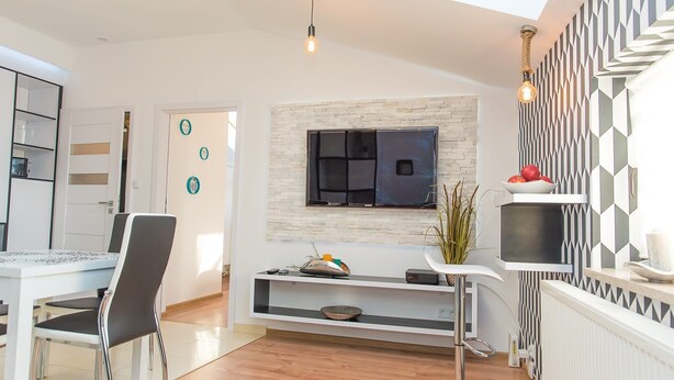 A modern living room with a flat-screen TV mounted on a stone wall, a dining area with a white table and chairs, and a decorative wall with geometric patterns.
