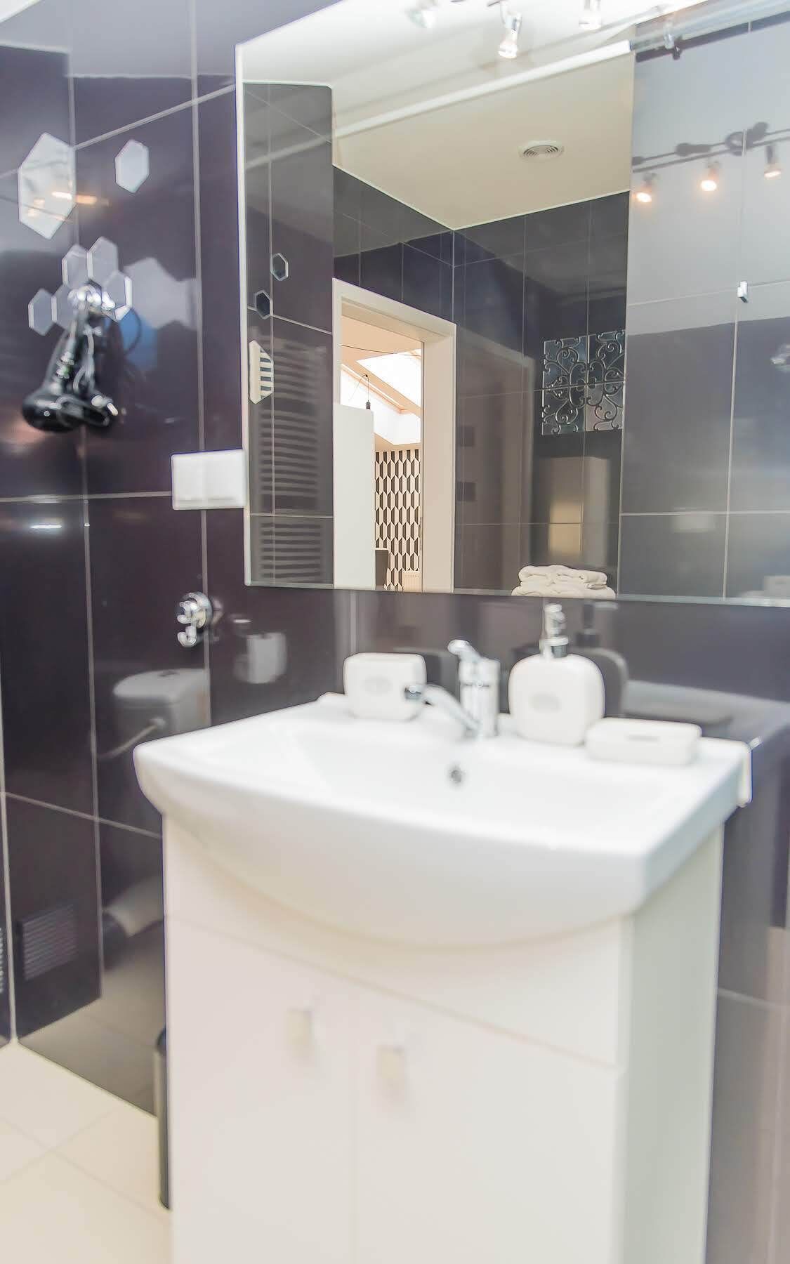 Un baño moderno con un espejo grande, lavamanos blanco y paredes de azulejos oscuros.