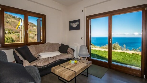 A living room with a sofa, a coffee table, and a view of the sea through large sliding glass doors. - Villa Mylopotamos (Zagora-Mouresi)
