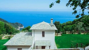 A white building with a stone roof, surrounded by a green lawn and a view of the sea. - Villa Mylopotamos (Zagora-Mouresi)