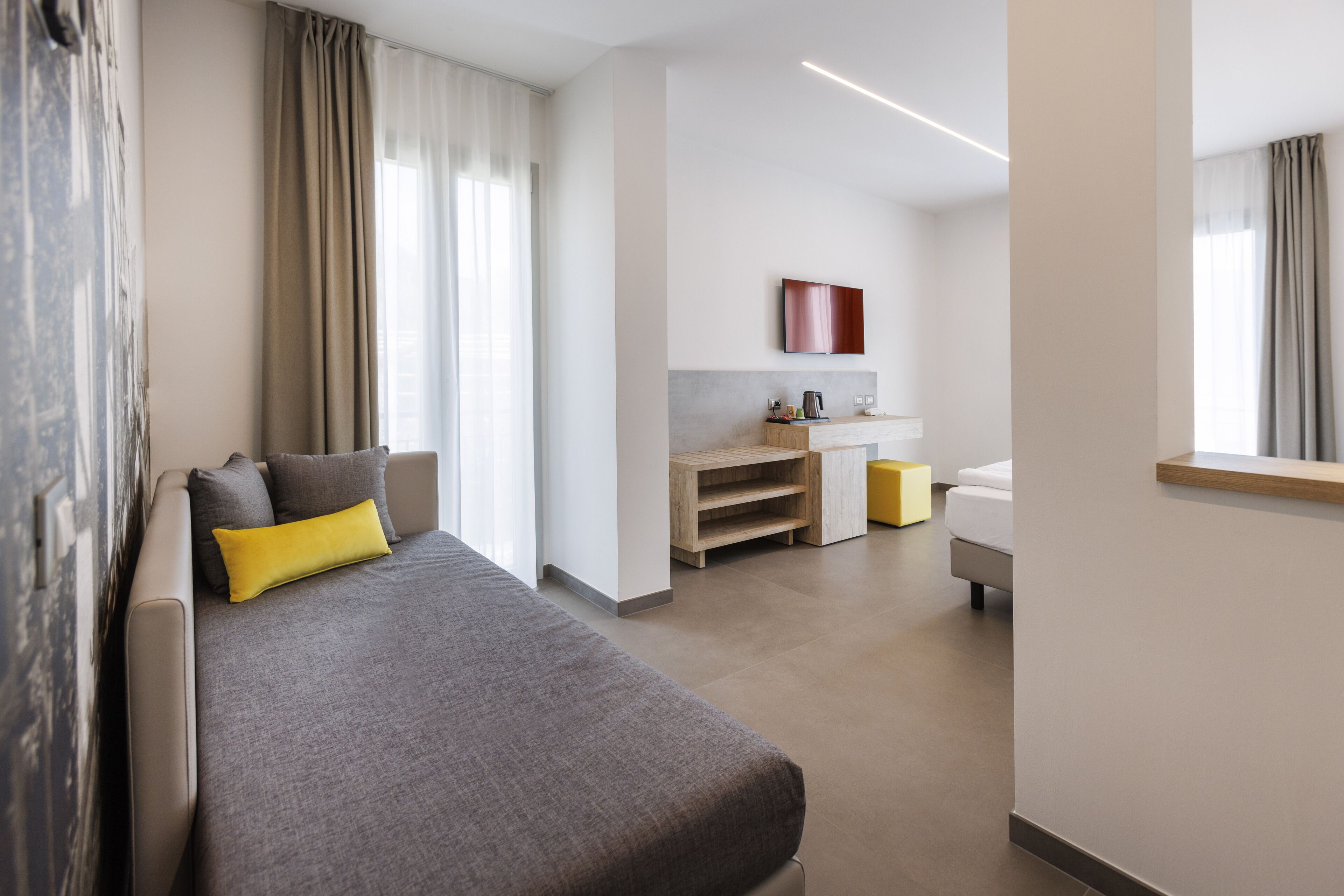 A modern hotel room with a grey sofa, a small wooden side table, a yellow ottoman, a bed, a bathroom, and a kitchen area.