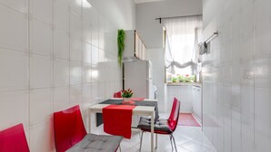 A compact kitchen with white tiled walls, a dining table with red chairs, and a small window with curtains.