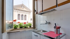 A small kitchen with white cabinets, a window with potted plants, and a view of a building with arched windows.
