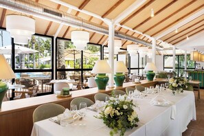 Un restaurant avec des tables dressées pour le service, de grandes fenêtres et des plantes d’intérieur.