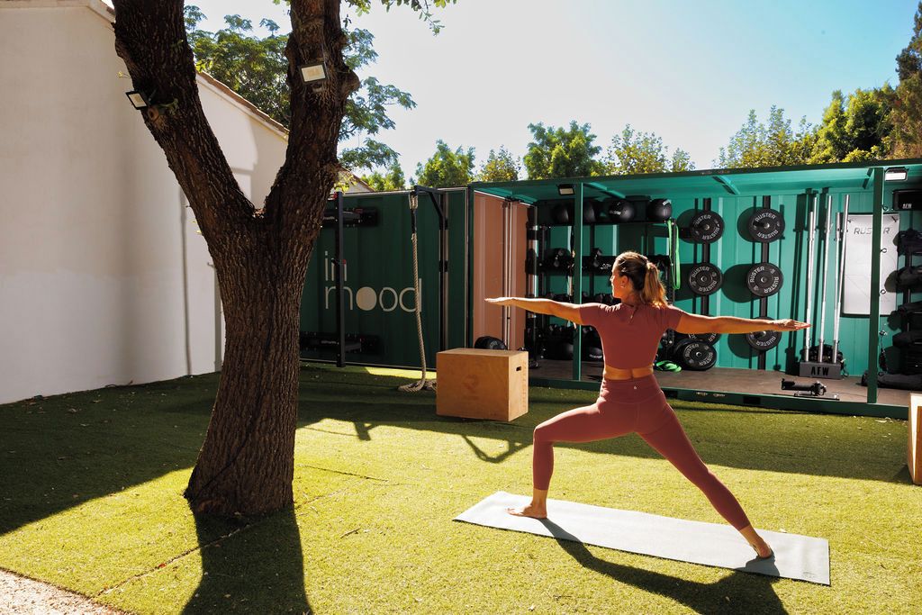 Una persona realizando una postura de yoga sobre una esterilla al aire libre, con un árbol y una estructura verde al fondo.