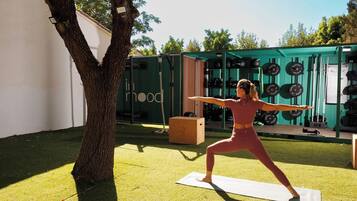 Una persona realizando una postura de yoga sobre una esterilla al aire libre, con un árbol y una estructura verde al fondo.