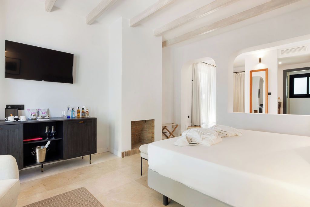 A modern bedroom with a large bed, a flat-screen TV mounted on the wall, a dark wooden dresser, and a fireplace.